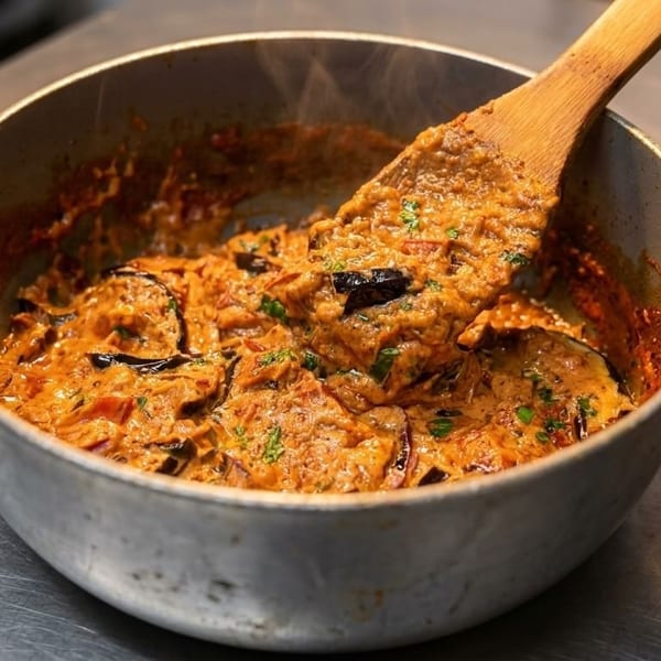 A wooden spoon stirs a creamy, orange-colored dish with eggplant slices in a metal pan.